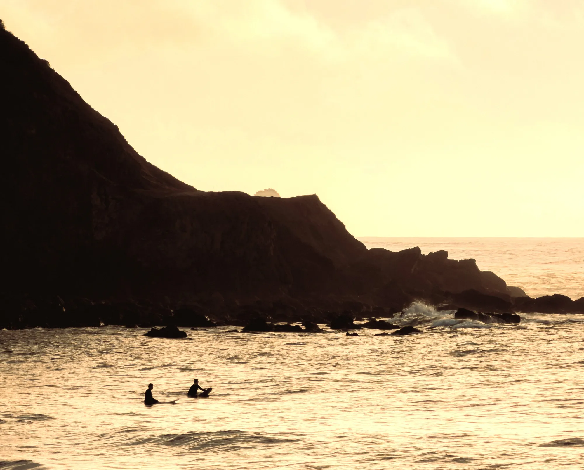 two people sitting on surfboards on the sea with mountains in the back